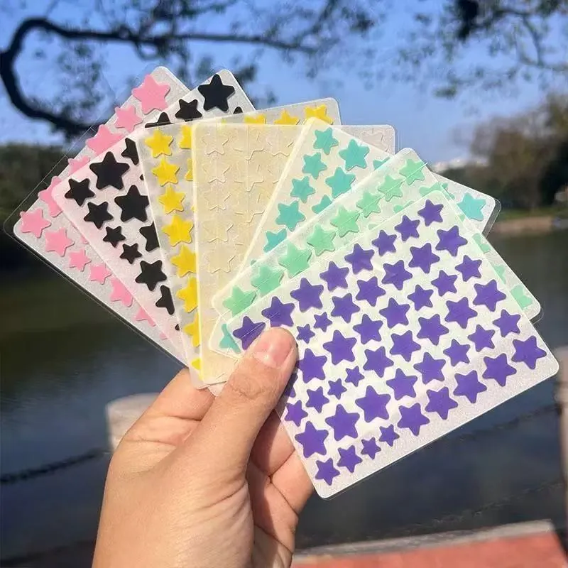 Mão segurando cartelas de adesivos em forma de estrela nas cores rosa, preta, amarela, creme, verde e roxa, ao ar livre