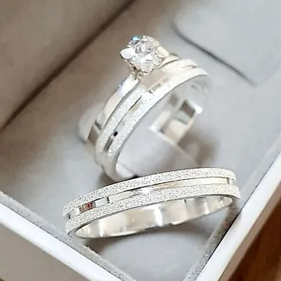 Duas alianças prateadas com faixas jateadas e friso liso e um anel solitário com pedra clara numa caixa cinza