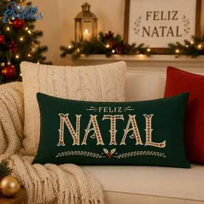 Almofada retangular verde-escura com Feliz Natal em letras creme e azevinho, sobre sofá claro com manta de tricô