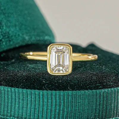 Anel dourado de aro fino com pedra retangular transparente em moldura fechada, sobre caixa de veludo verde