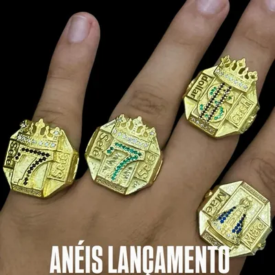 Mão com quatro anéis dedeira dourados: dois com número 7 e coroa, um com cifrão e um com Nossa Senhora em pedras azuis