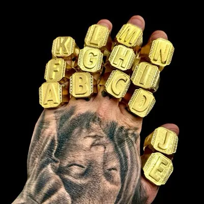 Mão tatuada usando vários anéis dourados de letra, de A a N, com moldura de pontos brilhantes, em fundo preto