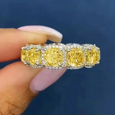 Anel prateado com zircônias amarelas quadradas cercadas por pedrinhas claras, seguro entre dedos sobre fundo azul