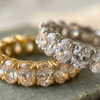 Dois anéis eternidade com zircônias ovais, um dourado e um prateado, apoiados sobre uma pedra esverdeada