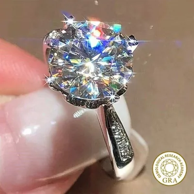 Anel prateado com grande moissanite redonda em garras de coroa e pedrinhas no aro, segurado entre os dedos