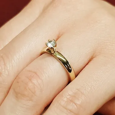 Anel solitário dourado com aro liso e pedra clara presa em garras, usado no dedo anelar de uma mão feminina