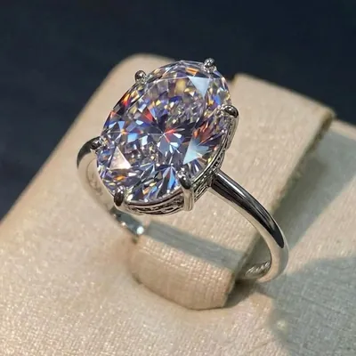 Anel prateado com grande zircônia oval presa por garras, encaixado numa caixinha de anel de tecido bege