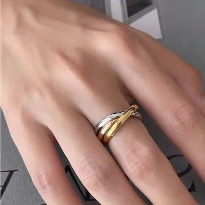 Mão feminina usando anel de três aros entrelaçados nos tons prateado, dourado e rosé sobre fundo cinza