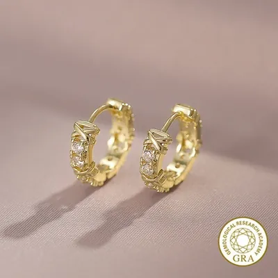 Par de argolinhas douradas com pedrinhas claras em pares alternadas com detalhes em X, sobre fundo rosado