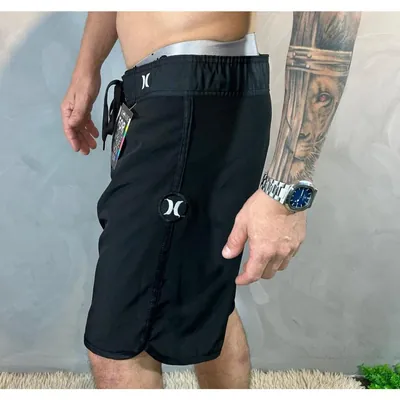 Homem de perfil vestindo bermuda surf preta na altura do joelho, com cordão na cintura, logotipo branco e barra arredondada