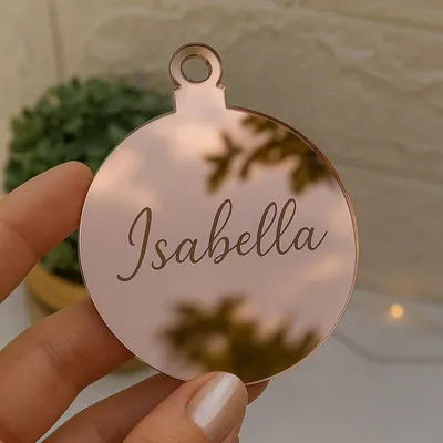 Mão segurando enfeite plano em forma de bola de Natal espelhada rosé com o nome Isabella gravado em letra cursiva