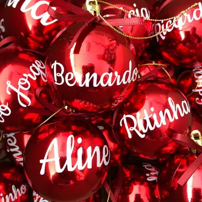 Várias bolas de Natal vermelhas brilhantes com nomes escritos em letra cursiva branca, laços de cetim e cordão dourado