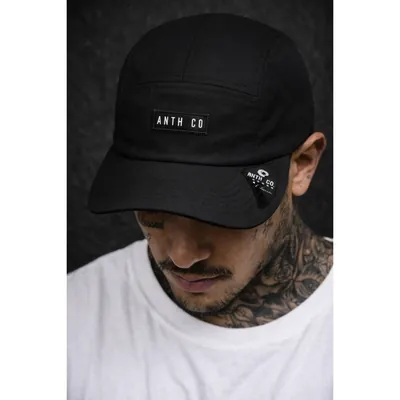 Homem tatuado de camiseta branca usando boné five panel preto de aba curva com etiqueta na frente e tag na aba