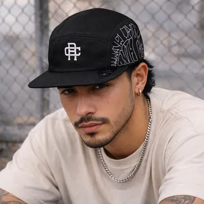 Rapaz de camiseta bege e corrente prateada usa boné five panel preto com monograma branco, em frente a uma grade