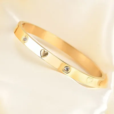 Bracelete rígido dourado e polido com coração em relevo, coração gravado e pedrinhas redondas, sobre tecido acetinado claro