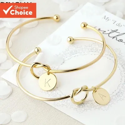 Dois braceletes dourados abertos com nó no aro e plaquinhas redondas com as letras K e A sobre cartão de casamento