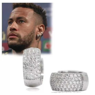 Par de argolas largas cor prata cravejadas de zircônias e, ao fundo, foto de jogador de futebol usando brincos