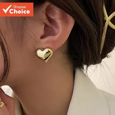Orelha com brinco de coração dourado estufado e polido; modelo de blazer branco e cabelo preso com piranha dourada