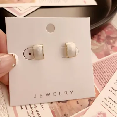Par de brincos pequenos de meia argola em esmalte branco perolado com borda dourada, presos num cartão branco