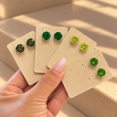 Mão segurando quatro cartelas com brincos ponto de luz em tons de verde: oliva, esmeralda, verde-limão e verde vivo