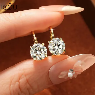 Dedos com unhas decoradas segurando par de brincos dourados com moissanite redonda presa por quatro garras