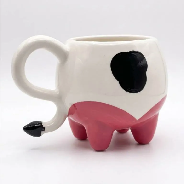 Caneca branca em formato de vaca com mancha preta, base rosa de úbere apoiada nas tetas e alça em forma de rabo