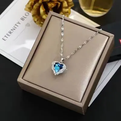Colar prateado com pingente de coração de cristal azul e moldura de pedrinhas, apoiado numa caixa bege