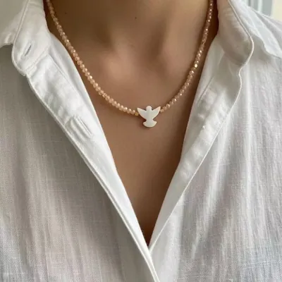 Colar de continhas cor pêssego com pingente branco em forma de pomba no pescoço de mulher de camisa branca de linho
