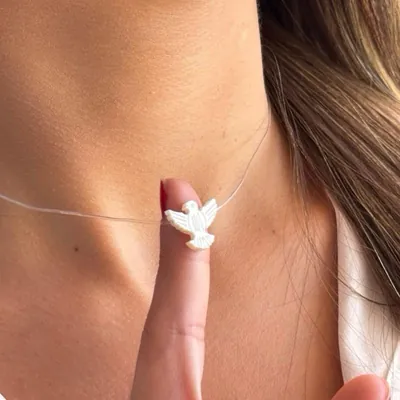 Pingente branco de pombinha do Espírito Santo em madrepérola num fio transparente, apoiado na ponta do dedo junto ao pescoço