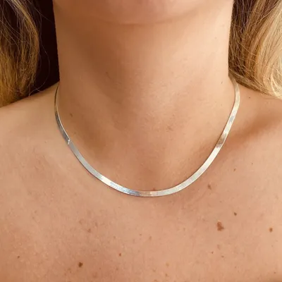 Colar de prata chato e laminado, tipo fita, usado rente à base do pescoço por mulher de cabelo loiro