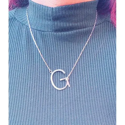Colar prateado com letra G grande presa de lado na corrente, usado sobre blusa canelada verde-escura de gola alta