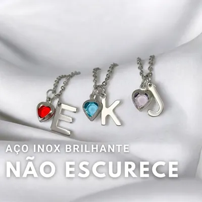 Três colares prateados com letras E, K e J e corações de pedra vermelha, azul e lilás sobre tecido branco acetinado