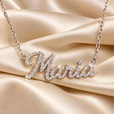 Colar prateado com o nome Maria em letra cursiva cravejada de pedrinhas, sobre tecido acetinado bege