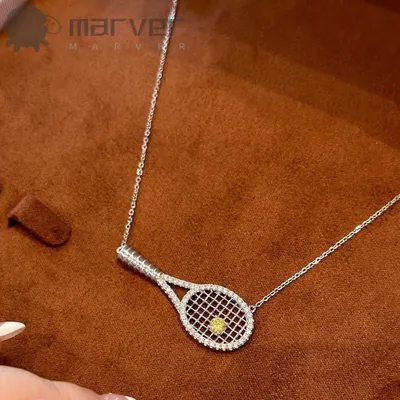 Colar prateado com pingente de raquete de tênis cravejada e pedra amarela no centro, sobre camurça marrom