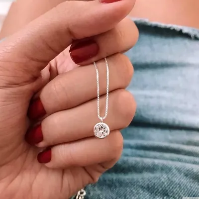Mão de unhas vermelhas segurando colar prateado fino com pingente redondo de pedra clara em aro