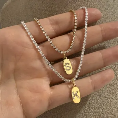 Dois colares riviera na palma da mão, um dourado e um prateado, com plaquinhas douradas das letras S e K cravejadas