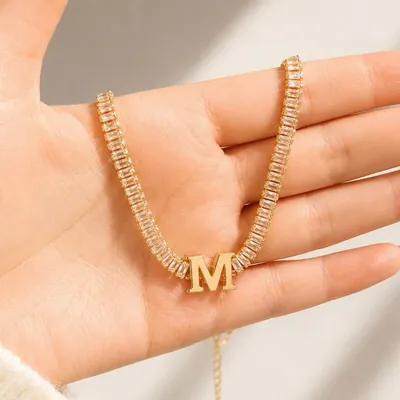 Mão segurando colar riviera dourado de zircônias retangulares com letra M lisa no centro