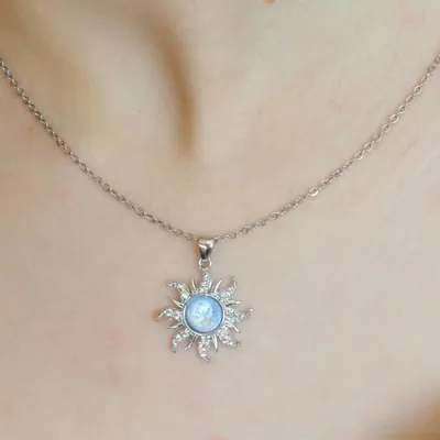 Pingente prateado em forma de sol com raios ondulados e pedra azul-clara no centro, em corrente fina no pescoço