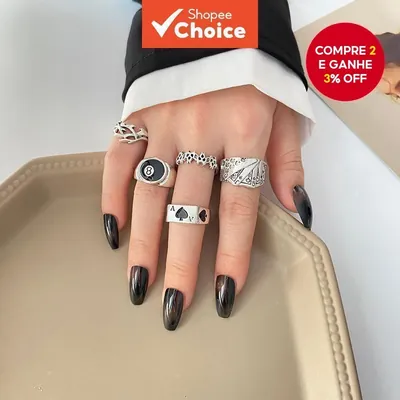 Mão com unhas pretas usando cinco anéis prateados: espinhos, bola 8, chamas, cartas de baralho e ás de espadas