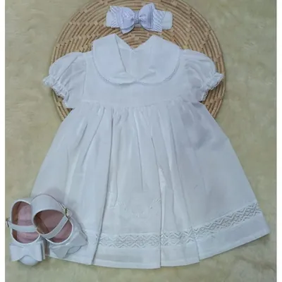 Vestido branco de batizado com gola de renda, faixa de laço e sapatinhos brancos de verniz sobre jogo americano de palha