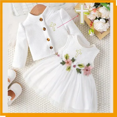 Vestido branco de bebê com saia de tule, flores bordadas e cruz dourada ao lado de cardigã branco de manga longa