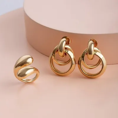 Anel dourado aberto com pontas em gota e par de brincos com duas argolas entrelaçadas, sobre fundo rosado
