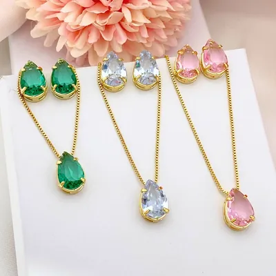 Três conjuntos de colar e brincos em gota, verde, azul-claro e rosa, com correntes douradas e flor rosa ao fundo