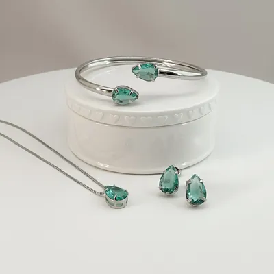 Conjunto prateado com pedras gota verdes: bracelete aberto sobre caixinha branca, colar e par de brincos