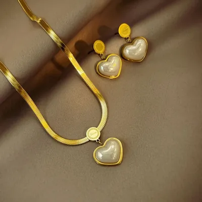 Colar dourado de corrente achatada com pingente de coração perolado e par de brincos iguais sobre tecido acetinado marrom