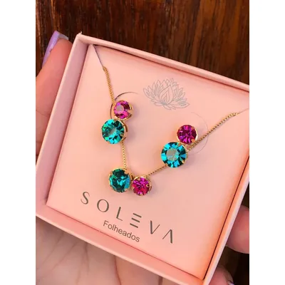 Colar e par de brincos dourados com pedras redondas pink e verde-azuladas em caixa rosa segurada na mão
