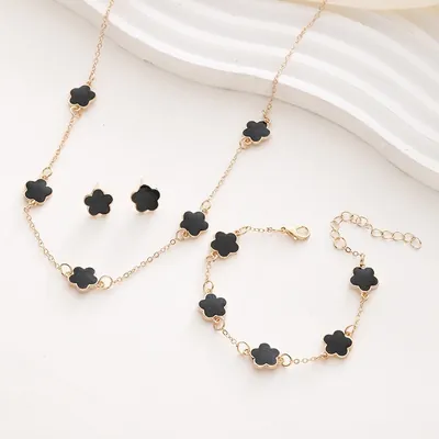 Colar, pulseira e par de brincos dourados com flores pretas de cinco pétalas sobre superfície branca ondulada