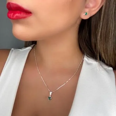 Mulher de batom vermelho usando corrente fina com pingente de coração verde e brinco redondo verde
