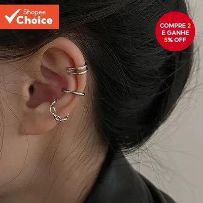 Orelha com três ear cuffs prateados na cartilagem: um duplo, um liso e um trançado, sobre cabelo escuro preso