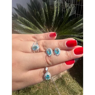Mão de unhas vermelhas com anel, par de brincos e pingente com pedra oval azul cercada de pedrinhas brancas